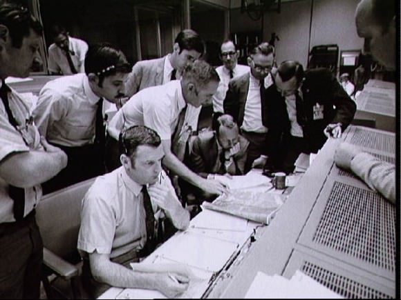 Flight controllers and managers gather around the console of Flight Director Glynn Lunney (seated in white shirt, foreground). At far left, in the dark suit, is Chris Kraft, deputy director of the Manned Spacecraft Center. Credit: NASA.