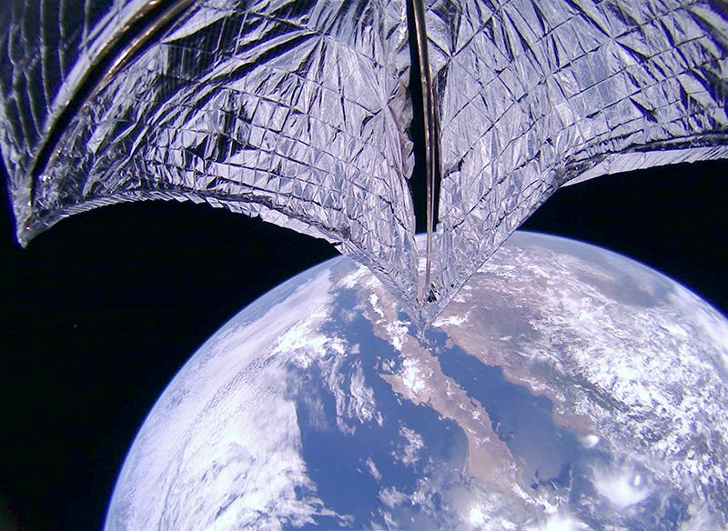 A dramatic image of the sails being deployed, with you-know-what in the background. Image Credit: The Planetary Society.