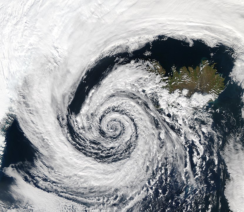 This is an image near Iceland. Air is rushing in to fill an area of low atmospheric pressure. As the air moves, it's subject to the coriolis force, producing the spiral. Image Credit: By NASA’s Aqua/MODIS satellite - https://visibleearth.nasa.gov/view.php?id=68992, Public Domain, https://commons.wikimedia.org/w/index.php?curid=400656