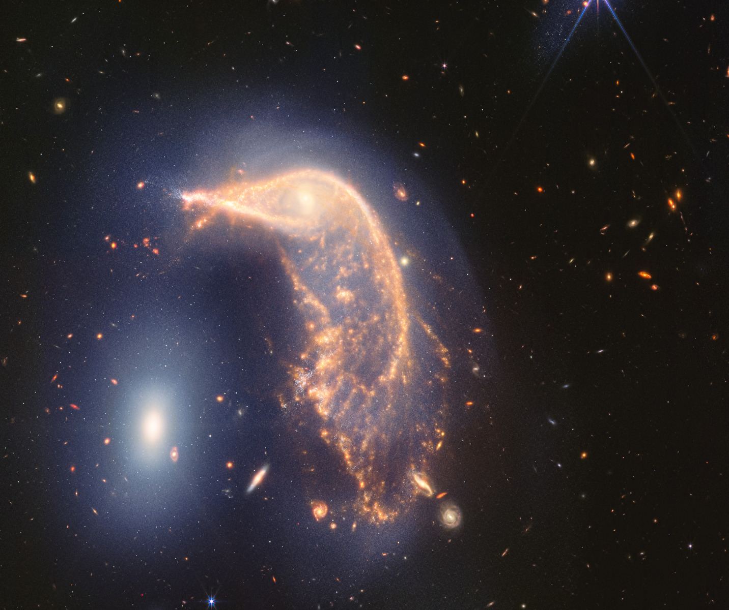 This “penguin party” (called Arp 142) is loud! The distorted spiral galaxy at center, the Penguin, and the compact elliptical galaxy at left, the Egg, are locked in an active embrace. A new near- and mid-infrared image from the Webb Space Telescope, taken to mark its second year of science, shows that their interaction is marked by a faint upside-down U-shaped blue glow. The blue galaxy at upper right (near bright star) is a closer galaxie teeming with new stars. It's not part of the collision and lies closer to Earth than Arp 142.